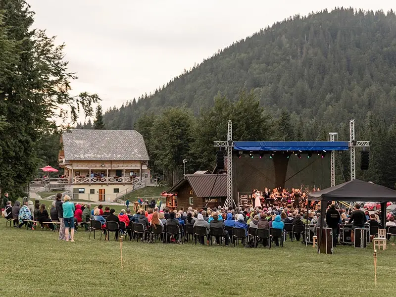 {"sl":"Skupina ljudi sedi na travniku pred odprtim odrom, obdanem z gozdom in hribi.","en":""}